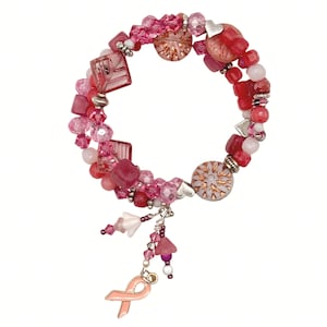 May include: A multi-strand bracelet featuring a variety of pink and red beads, including faceted crystals, smooth round stones, and square-shaped glass pieces. The bracelet is accented with small silver heart charms and a dangling pink awareness ribbon.