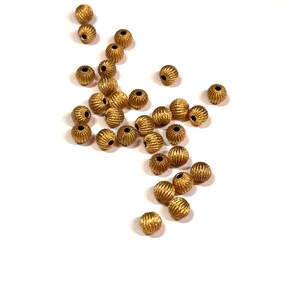 May include: A scattering of small, gold-coloured beads on a white surface. Each bead features a textured, ridged surface and a small hole. These decorative beads are suitable for jewellery making and craft projects.
