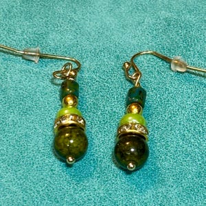 Puede incluir: Un par de pendientes colgantes con ganchos de metal dorado. Cada pendiente consta de una cuenta cilíndrica azul verdoso, un pequeño espaciador dorado, una cuenta redonda verde lima, un anillo con incrustaciones de pedrería y una cuenta verde oliva.