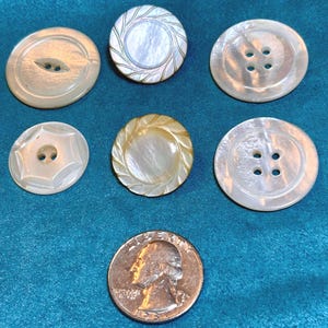 May include: A collection of six round, iridescent mother-of-pearl buttons in various designs, including two-hole and four-hole styles, arranged alongside a United States quarter coin for scale, showcasing natural white and cream color variations.