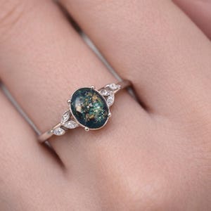 May include: A silver ring featuring an oval-shaped, dark teal gemstone with flecks of green and gold. The ring is accented with small, clear gemstones and leaf-shaped details on the band, creating an elegant design.