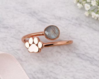 Puede incluir: Un anillo de oro rosa con un diseño de huella de pata y una gema gris redonda. El anillo presenta un diseño envolvente con la huella de la pata en un extremo y la gema en el otro. El anillo está sobre un fondo de mármol.