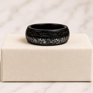 May include: A black ring featuring a textured, dark surface with a horizontal band of shimmering, metallic-like silver and grey flecks embedded around the center, creating a contrast against the smooth, matte black finish of the band's edges.