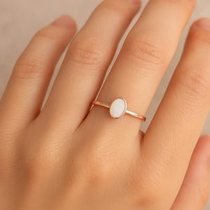 May include: A rose gold ring featuring a white oval stone. The ring is simple and elegant, with a thin band and a bezel setting for the stone. The ring is shown on a hand.
