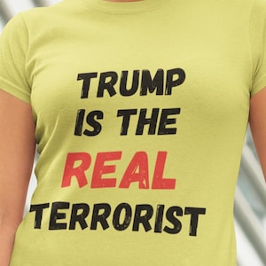 May include: A light green t-shirt with the text "TRUMP IS THE REAL TERRORIST" in black and red. The shirt is a crew neck style and is being worn.