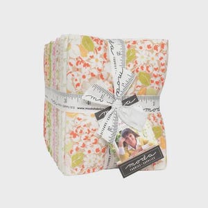 May include: A stack of floral fabric squares tied with a white ribbon featuring a ruler design. The fabric has a pattern of white flowers, orange and green leaves on a beige background. A tag with a woman's photo and the words "moda FABRICS + SUPPLIES" is attached.