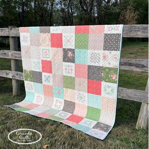 May include: A patchwork quilt with a variety of square patterns in shades of pink, coral, green, gray, and blue. The quilt is draped over a wooden fence. The quilt has a light blue border and the text "Coriander Quilts" in a white oval.