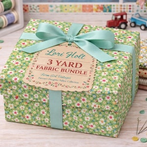 May include: A green floral fabric bundle tied with a light blue ribbon. The tag reads "Lori Holt 3 Yard Fabric Bundle." The fabric is a light green with small white and yellow flowers. Sewing notions and spools of thread are in the background.