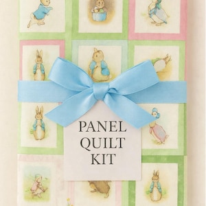 May include: A Panel Quilt Kit featuring illustrations of Peter Rabbit and friends. The fabric is arranged in a grid pattern with green and pink borders. A light blue ribbon and bow wrap the kit, with a tag that reads "Panel Quilt Kit."