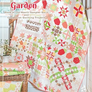 May include: A colorful quilt with a strawberry theme, featuring red, green, and peach-colored patterns. The quilt is draped over a railing, with a stack of folded quilts and a basket in the foreground. The text "Strawberry Garden" is visible.