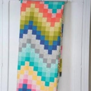 May include: A colorful woven blanket with a geometric pattern of squares in shades of pink, orange, yellow, green, blue, gray, and teal. The blanket is hanging on a metal hanger against a white door.