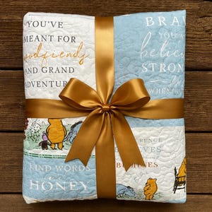 May include: A light blue and white quilt with a gold ribbon and bow. The quilt features Winnie the Pooh illustrations and inspirational text. The text includes phrases like "You've meant for good friendly and grand adventure."