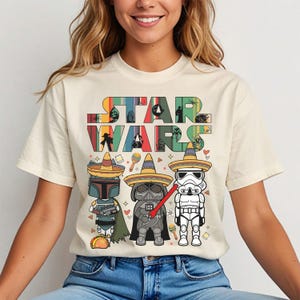 Könnte beinhalten: Ein cremefarbenes Kurzarm-T-Shirt mit dem Schriftzug STAR WARS in einer bunten, gestreiften Schriftart. Darunter tragen drei Zeichentrickfiguren Sombreros: ein Kopfgeldjäger, Darth Vader mit einem roten Lichtschwert und ein Sturmtruppler, ergänzt durch Taco- und Maraca-Symbole.
