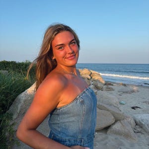 May include: A woman wearing a strapless denim dress stands on a sandy beach with the ocean in the background.