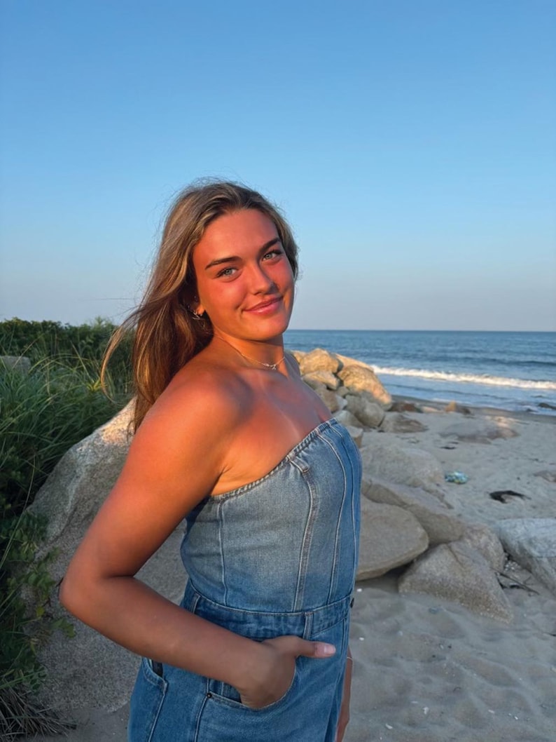 May include: A woman wearing a strapless denim dress stands on a sandy beach with the ocean in the background.