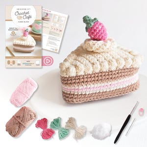 May include: A crochet kit for a Cake Slice, featuring a large, handmade slice of cake with a strawberry on top. The kit includes yarn, a crochet hook, and instructions. The packaging reads "Crochet Café - Cake Slice" and "Great for Beginners!"