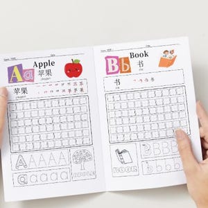 May include: Open workbook pages with the words "Apple" and "Book" in English and Chinese. The "Apple" page has a red apple illustration. The "Book" page has a small illustration of a book and people reading. The workbook pages have lined boxes for writing practice.