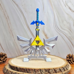 Puede incluir: Una réplica impresa en 3D de una espada y un emblema. La espada presenta una empuñadura azul con una hoja gris y un emblema triangular amarillo. El emblema es un diseño alado plateado con un triángulo amarillo central sobre una base.