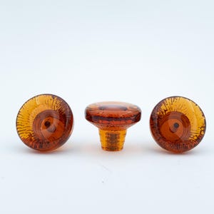 May include: Three amber-coloured glass cabinet knobs with a circular, translucent design. Two knobs are positioned to show their flat, patterned faces, while the centre knob is turned to display its side profile and threaded metal attachment.
