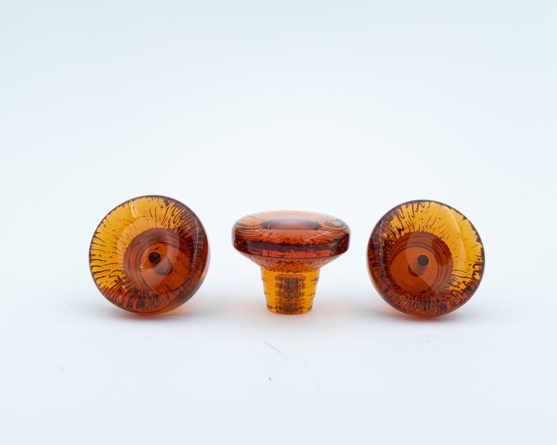 May include: Three amber-coloured glass cabinet knobs with a circular, translucent design. Two knobs are positioned to show their flat, patterned faces, while the centre knob is turned to display its side profile and threaded metal attachment.