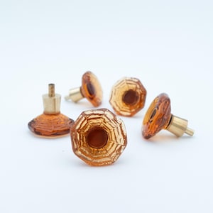 Op de afbeelding: Een verzameling van vijf amberkleurige glazen kastknoppen met messing hardware. De knoppen hebben een facet, geometrisch ontwerp en zijn gerangschikt op een witte ondergrond. Geschikt voor meubels.
