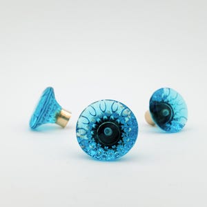 Könnte beinhalten: Drei dekorative Schrankknöpfe aus türkisfarbenem Glas. Die Knöpfe haben ein strukturiertes, blumenartiges Design und einen goldfarbenen Sockel. Die Knöpfe sind auf weißem Hintergrund angeordnet.
