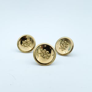 May include: Three gold-coloured, round cabinet knobs with a decorative swirl design in the centre. The knobs are made of a shiny, reflective metal against a white background.