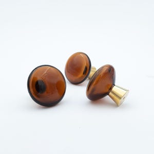 May include: Three round cabinet knobs featuring translucent amber-coloured glass tops with a smooth, polished finish. Each knob is attached to a conical, metallic gold-toned base that tapers towards the centre, providing a sturdy mounting point.