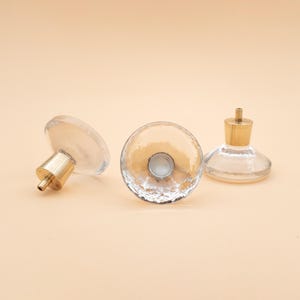May include: Three clear glass bottle stoppers with gold-colored metal accents are arranged on a light peach background. The stoppers have a round, textured design and a flat base.