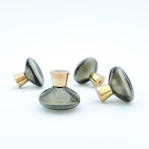 May include: Four decorative glass knobs with brass accents. The knobs are a smoky gray color with a rounded shape. The brass accents are a warm gold color. These knobs are ideal for furniture or cabinet upgrades.
