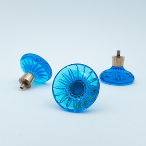 Puede incluir: Tres pomos de cristal azul brillante con herrajes de latón. Los pomos tienen un diseño facetado en forma de estrella. Estos pomos son probablemente para muebles o fines decorativos. El fondo es blanco liso.