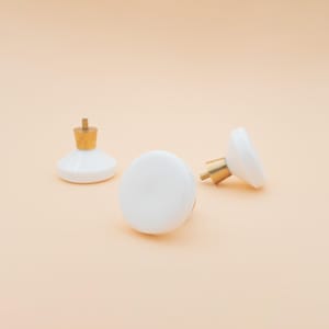 Könnte beinhalten: Drei weiße Keramik-Schrankknöpfe mit Messingbeschlägen. Ein Knopf steht aufrecht, zwei liegen auf der Seite. Die Knöpfe haben ein schlichtes, rundes Design, und die Messingbeschläge verleihen einen Hauch von Eleganz.