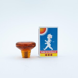 May include: An amber-coloured glass knob with a circular, faceted top and a tapered base, positioned next to a rectangular matchbox featuring a blue background, a white silhouette of a child, a yellow sun, and the text SOLSTICKAN with three red stars.