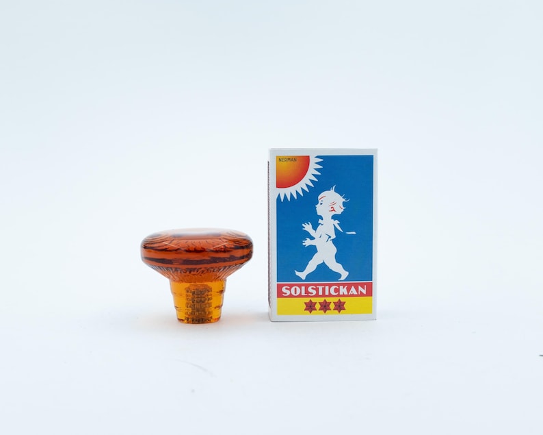 May include: An amber-coloured glass knob with a circular, faceted top and a tapered base, positioned next to a rectangular matchbox featuring a blue background, a white silhouette of a child, a yellow sun, and the text SOLSTICKAN with three red stars.