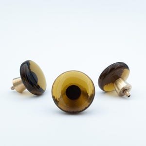 Puede incluir: Tres pomos decorativos para armarios con un diseño redondo de cristal color ámbar y un acento marrón oscuro. Cada pomo tiene una base de metal color latón. Los pomos están dispuestos sobre un fondo blanco.