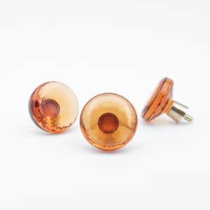 May include: Three amber glass cabinet knobs with a round, slightly raised center. One knob has a metal post attached to the back.