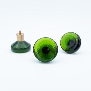May include: Three green glass objects on a white background. One is a small, round bottle with a gold-colored stopper. Two are round, green glass spheres. The glass has a textured surface and a dark center.