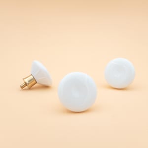 May include: Three white ceramic cabinet knobs with brass hardware. One knob is angled, showing the brass screw. The other two knobs are round and smooth. The knobs are on a light peach background.