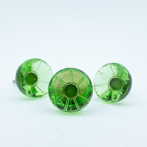 May include: Three round, translucent green glass cabinet knobs featuring a radial, sunburst-like pattern of raised lines extending from a central circular indentation. Each knob has a smooth, domed surface and a metallic mounting screw visible on the side.
