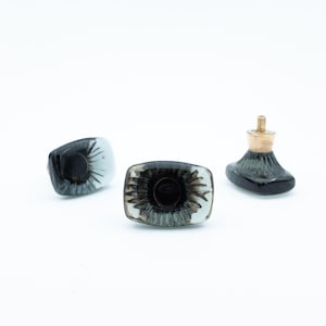 May include: Three rectangular cabinet knobs with a clear glass front featuring a black iris and pupil design that resembles a human eye. Each knob has a metallic gold-toned base and a short cylindrical stem for mounting to furniture surfaces.