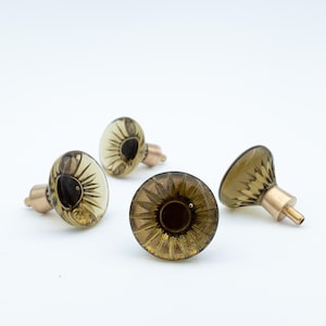 Peut inclure: Quatre boutons de meuble décoratifs. Les boutons présentent un verre transparent de couleur ambre avec un motif en forme d'éclat de soleil et une base de couleur laiton. Les boutons sont disposés sur un fond blanc.