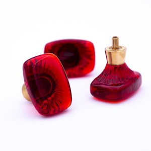 Puede incluir: Tres tapones decorativos para botellas. Dos son rectangulares con detalles en latón, y uno tiene una forma única con una parte superior dorada. Todos son de color rojo intenso y transparente.