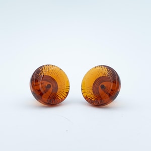 May include: A pair of round, amber-coloured glass stud earrings featuring a translucent, swirling pattern of dark brown lines that radiate from a central point, creating a distinct, layered effect within the smooth, polished surface of each piece.