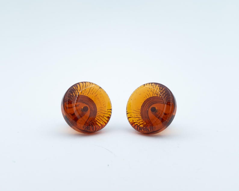 May include: A pair of round, amber-coloured glass stud earrings featuring a translucent, swirling pattern of dark brown lines that radiate from a central point, creating a distinct, layered effect within the smooth, polished surface of each piece.