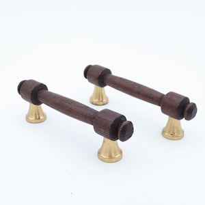 Peut inclure: Deux poignées de tiroir composées d'une barre horizontale en bois brun foncé aux extrémités arrondies et de deux bases coniques en métal doré poli. Chaque poignée présente une section centrale cylindrique en bois avec des accents circulaires.