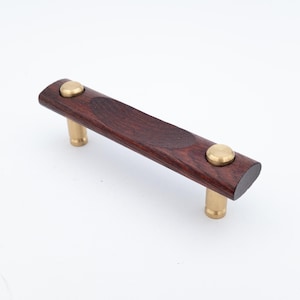 Peut inclure: Une poignée de meuble en bois foncé avec des éléments en laiton. La poignée est d'un brun rougeâtre riche avec un design légèrement incurvé. Les éléments en laiton comprennent deux accents ronds et deux montants cylindriques.