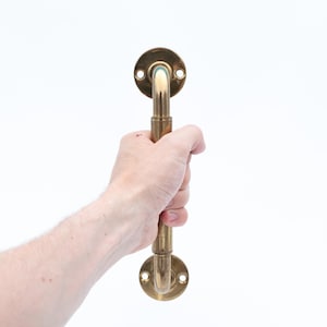 Può includere: Una maniglia per porta in ottone con una piastra di montaggio circolare ad ogni estremità. La maniglia è tenuta in una mano su sfondo bianco. La maniglia ha un design curvo ed è realizzata in ottone.