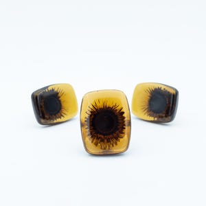 May include: Three rectangular buttons with rounded corners, featuring a translucent amber-colored resin body. Each button contains a dark brown, sunburst-like floral pattern embedded in the center, creating a layered, depth-filled appearance.