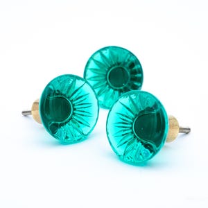 Op de afbeelding: Drie teal glazen kastknoppen met een decoratief sunburst ontwerp. Elke knop heeft een messingkleurige basis. De knoppen zijn gerangschikt op een witte achtergrond.