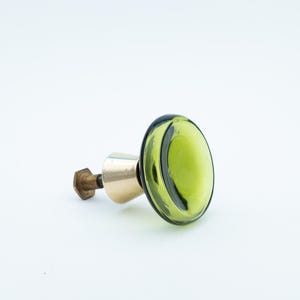 Pode incluir: Um puxador decorativo para armário com uma face redonda de vidro verde-oliva. O puxador tem uma base de cor latão e um parafuso de cor bronze. O vidro tem uma borda verde escura. O puxador é projetado para móveis e decoração de casa.